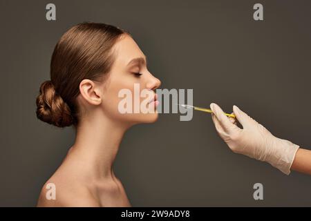 esthéticienne en gant médical tenant la seringue près de la jeune femme sur fond gris, vue de côté Banque D'Images