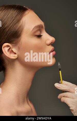 esthéticienne en gant médical tenant la seringue près des lèvres de la jeune femme sur fond gris, vue de côté Banque D'Images