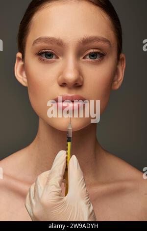 esthéticienne en gant médical tenant la seringue près de la jeune femme sur fond gris, remplisseur de lèvres Banque D'Images
