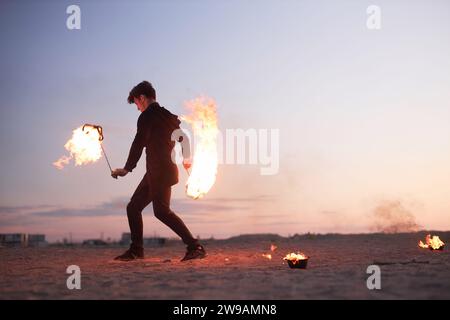 Vue latérale pleine longueur de l'artiste de spectacle de feu masculin dansant avec des flammes dans la nature au coucher du soleil, copie espace Banque D'Images