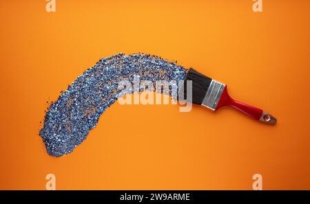 Un pinceau large dessine un coup de pinceau avec des paillettes brillantes bleu vif sur fond orange. Banque D'Images