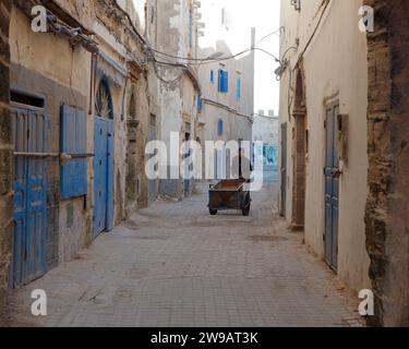 Un homme âgé pousse un chariot en bois le long d'une rue poussiéreuse dans la médina avec des portes et des volets en bois bleu à Essaouira, Maroc. 26 décembre 2023 Banque D'Images