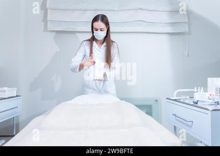 Le médecin se prépare à recevoir un patient dans un salon de beauté. La femme couvre le canapé avec une couverture blanche hygiénique protectrice. Supports mixtes Banque D'Images