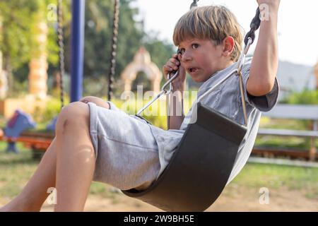 Garçon de 6 à 7 ans en T-shirt en coton gris est assis sur la balançoire dans l'aire de jeux pour enfants et a peur. Banque D'Images