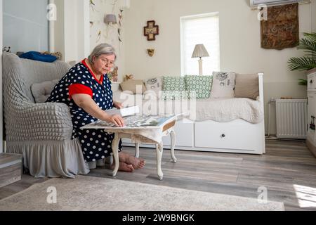 Une femme âgée collectionnant le puzzle tout en étant assise dans une chaise dans sa chambre lumineuse à la maison. Banque D'Images