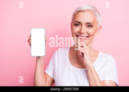 Photo de femme âgée intelligente avec coiffure courte porter blanc t-shirt touch menton montrer écran de smartphone isolé sur fond rose Banque D'Images