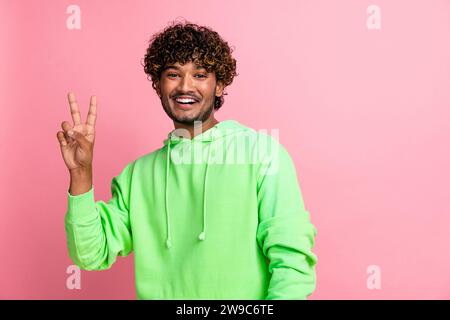 Portrait de joyeux jeune joyeux petit ami aux cheveux bouclés indien porter vert sweat à capuche geste v signe isolé sur fond de couleur rose Banque D'Images