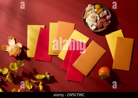 Pose à plat d'enveloppes porte-bonheur disposées au hasard sur un fond rouge avec une assiette de confiture, de mandarines et de branches jaunes de fleurs d'abricot. Scène activée Banque D'Images