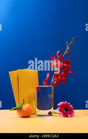 Une orchidée fraîche est placée dans un verre, deux enveloppes jaunes, une fleur de gerbera et une mandarine sont affichées sur une table beige avec un fond bleu. COP Banque D'Images