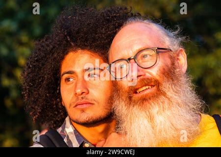 Dans cette image réconfortante, deux individus masculins, peut-être gays, partagent un moment de l’heure d’or. Leurs regards sont dirigés au loin, suggérant la contemplation ou une vision partagée. L'homme plus âgé, avec sa barbe blanche distinctive et ses lunettes, sourit largement, exsudant un sentiment de joie et de sagesse. Les cheveux bouclés et l'expression sincère de la jeune personne véhiculent un comportement réfléchi. La lumière chaude baigne leurs visages, soulignant les contrastes et les similitudes entre eux et créant une atmosphère de connexion intime. Compagnie Golden Hour. Photo de haute qualité Banque D'Images