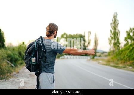 Jeune homme randonneur avec sac à dos sur l'auto-stop près de la route Banque D'Images