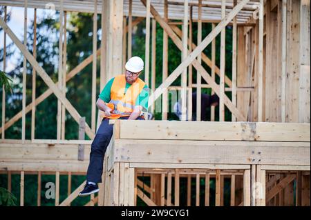 Menuisier construisant une maison à ossature en bois de deux étages près de la forêt. Homme barbu dans des lunettes martelant des clous dans la structure tout en portant un casque de protection. Concept de construction écologique moderne. Banque D'Images