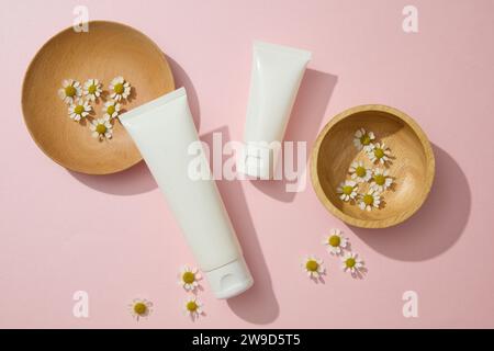 Fleurs de chamomilla fraîches décorées sur des plats en bois avec deux tubes en plastique récipient hydratant crème de nettoyant pour le visage sur un fond rose. Banque D'Images