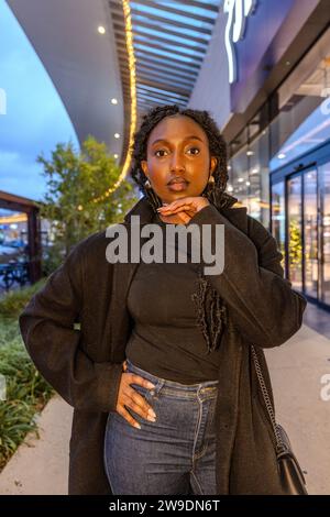 L'image capture un moment de réflexion d'une jeune femme noire avec sa main doucement posée sur son menton, comme si perdue dans les pensées, contre l'ambiance vibrante d'une avenue de détail urbaine animée. Sa tenue est un mélange de confort et de style avec un pardessus noir épais et un col roulé, serti contre le tissu denim doux de son Jean. Les plafonniers décoratifs créent un effet bokeh, donnant une touche fantaisiste au cadre urbain. Le regard ascendant de la femme et sa pose contemplative suggèrent un moment d'introspection au milieu de l'énergie de la vie citadine. Contemplative Dusk : une Rhapsodie de soirée urbaine. H Banque D'Images