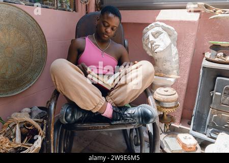 Femme afro haïtienne aux cheveux courts portant un pantalon en tissu brun et un haut rose, à la maison assise sur une chaise à bascule en bois lisant un livre roman, un concept de style de vie, Banque D'Images