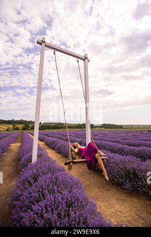 Belle jeune femme libre sur une balançoire parmi la lavande lumineuse et fraîche sur un champ. La beauté apprécie son humeur et la nature. Banque D'Images