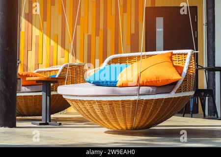 Oreillers colorés sur un lit de repos moderne. Chaises longues en rotin brun avec oreillers orange et bleu suspendus avec corde près de la terrasse relaxante. Transat pour bronzer Banque D'Images