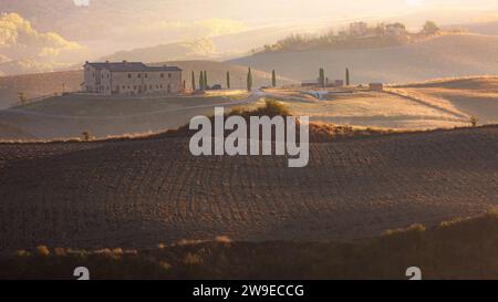 Lumière dorée matinale brumeuse au-dessus d'une maison toscane dans le paysage de campagne pittoresque de collines et de terres agricoles dans la Toscane rurale près d'Asciano, ITA Banque D'Images