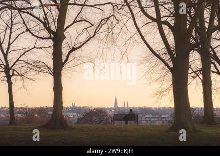 Moment de solitude comme une figure lointaine assis sur un banc de parc tranquille pendant un froid coucher de soleil ou lever de soleil d'hiver à Inverleith Park à Édimbourg, Écosse, Royaume-Uni Banque D'Images