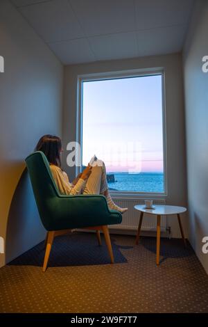 Femme en pyjama lisant dans une fenêtre face à la mer au coucher du soleil Banque D'Images