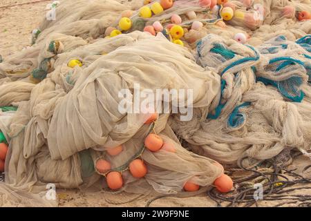 Une pile de filets de pêche colorés et de bouées se trouve sur une plage de sable. Les filets sont un mélange de bleus, verts et oranges, et les bouées sont rouges, jaunes et Banque D'Images