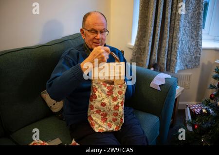 Homme retraité dans ses soixante-dix ans ouvrant un cadeau de noël tout en étant assis sur un canapé à côté de l'arbre de Noël dans une maison résidentielle, Angleterre, Royaume-Uni Banque D'Images