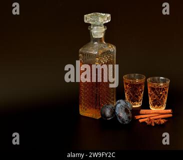 Liqueur de prune à la cannelle et à l'anis, alcool fort dans une carafe en cristal et deux verres sur fond noir, à côté d'épices et de fruits mûrs. Clos Banque D'Images