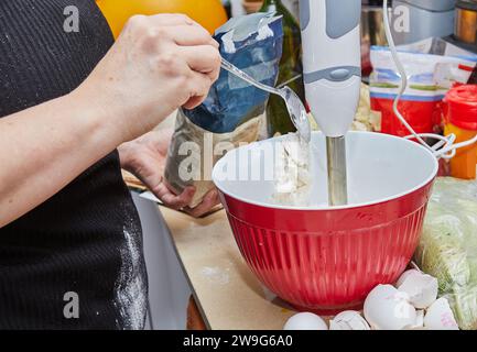 Une femme au foyer occupée est vue dans sa cuisine, fouettant habilement de la pâte à crêpes dans un bol à l'aide d'un mélangeur. L'image capture une scène domestique quotidienne o Banque D'Images