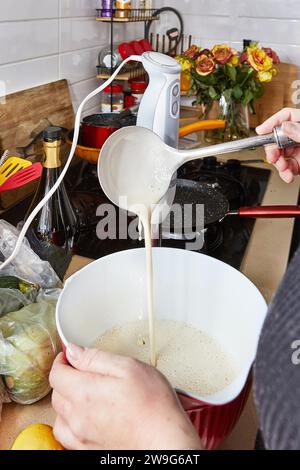 Gros plan d'une femme debout dans une cuisine rustique confortable versant un mélange de crêpes maison d'une louche dans un bol. Le mélange se déverse, créant un adorable Banque D'Images
