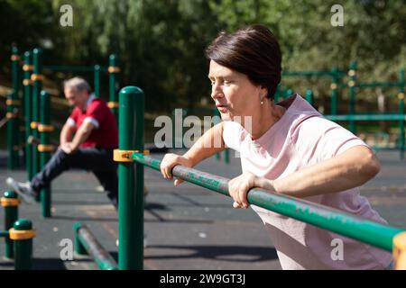 Femme âgée vigoureuse faisant des exercices du matin sur les barres de sport dans le terrain de sport en plein air. Homme faisant du sport en arrière-plan Banque D'Images