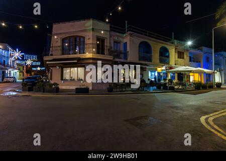 Un bâtiment d'angle illuminé avec un restaurant au niveau de la rue et une terrasse extérieure se dresse dans l'une des rues festivement éclairées de Marsaxlokk, Malte ; des guirlandes lumineuses et des installations lumineuses en forme d'étoile enjambent la rue déserte au premier plan. Banque D'Images