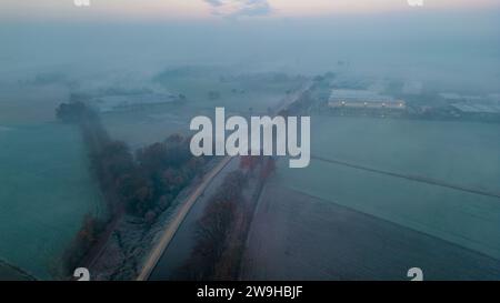 L'image présente une scène captivante tôt le matin où une brume dense couvre un paysage rural à l'aube. Une route droite traverse la scène, divisant les champs recouverts de gel qui affichent des nuances variables de bleus et de verts. La brume ajoute une couche de mystère et de douceur à l'environnement, obscurcissant partiellement les bâtiments et les arbres éloignés. Les tons de couleurs fraîches suggèrent le froid d'un matin d'hiver tôt. La qualité éthérée de la brume contraste avec la solidité de la route, créant un mélange d'éléments artificiels et naturels. L'atmosphère générale est calme et introspective, typique de t Banque D'Images