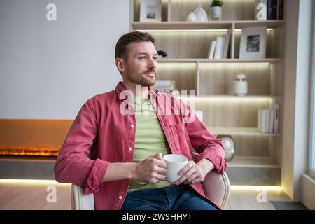 Homme positif calme assis dans un appartement moderne avec une tasse de thé, pensant, se reposant, regardant la fenêtre Banque D'Images