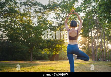 Jeune femme asiatique faisant du yoga le matin ou le soir au parc, femme en bonne santé se relaxant et pratiquant la pose d'arbre au parc de la ville. Pleine conscience, destinée, habitudes saines et concept d'équilibre Banque D'Images