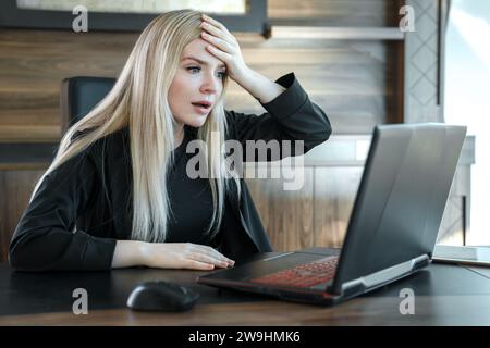 Femme avec l'expression effrayée regarde l'écran d'ordinateur portable, serrant son front avec sa main dans la surprise Banque D'Images