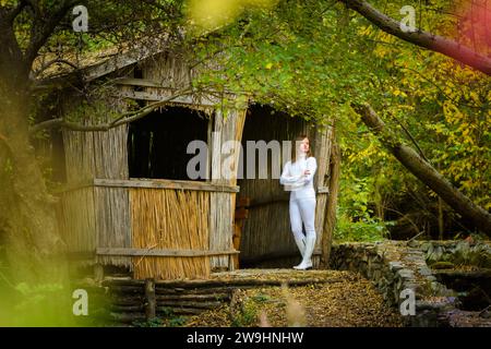 Une jeune belle fille en vêtements blancs se tient près d'une vieille maison abandonnée dans la forêt Banque D'Images