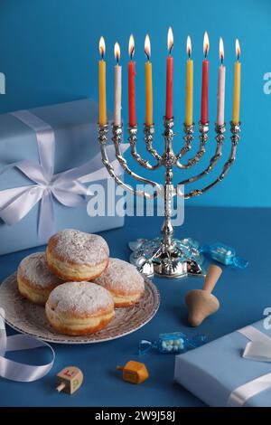 Célébration de Hanukkah. Menorah avec des bougies allumées, dreidels, beignets et boîte-cadeau sur la table bleu clair Banque D'Images