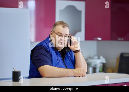 Conversation sérieuse au téléphone, grand homme caucasien dans ses années 40 avec des lunettes et les cheveux courts utilise smartphone dans la cuisine. Banque D'Images