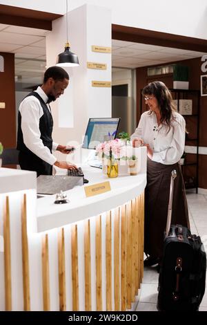 La réceptionniste afro-américaine aide la vieille femme à vérifier l'identité de l'hôtel et à récupérer les détails de réservation. Le concierge masculin tient et vérifie le passeport de la touriste féminine. Vue latérale. Banque D'Images