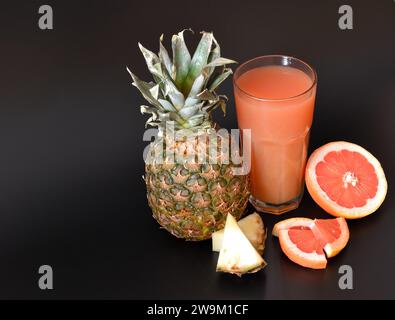 Un mélange de jus de fruits dans un grand verre sur fond noir, à côté de morceaux de pamplemousse mûr et d'ananas. Gros plan. Banque D'Images