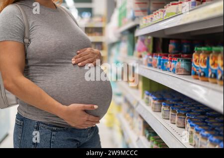La femme enceinte sans visage choisit la nourriture pour bébé dans le magasin. Pots avec purée de légumes. Banque D'Images