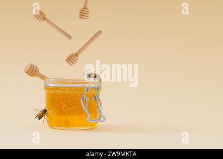 Mise en page créative faite avec des plongeurs de miel tombant dans un pot plein de miel naturel et de rayons de miel et des abeilles de miel volant autour sur fond crème clair. Banque D'Images