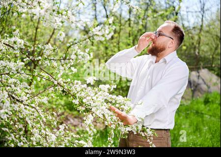 Homme allergique souffrant d'allergie saisonnière au printemps dans le jardin fleuri au printemps. Jeune homme avec des lunettes éternuant, fermant le nez à la main devant l'arbre en fleurs. Concept d'allergie printanière Banque D'Images