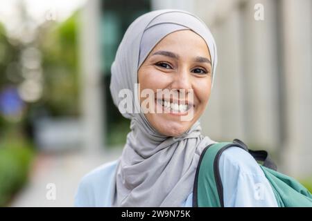 Portrait en gros plan d'une étudiante musulmane en hijab, femme avec un sac à dos derrière son dos souriant et regardant la caméra, debout à l'extérieur du campus du bâtiment universitaire. Banque D'Images