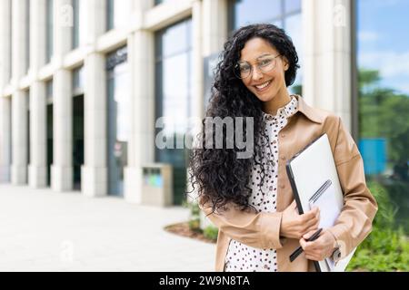 Portrait de jeune belle femme d'affaires réussie à l'extérieur de l'immeuble de bureaux, femme latino-américaine réussie souriant et regardant la caméra, debout avec l'ordinateur portable dans les mains et les documents. Banque D'Images
