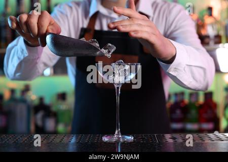 Préparation de cocktails alcoolisés. Barman ajoutant des glaçons dans le verre à Martini au comptoir dans le bar, gros plan Banque D'Images