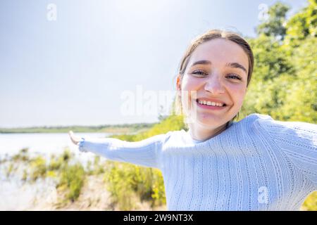 L'image capture une jeune femme joyeuse dans un chandail bleu clair, étendant son bras comme si elle montrait quelque chose au loin à un endroit pittoresque au bord du lac. Son sourire éclatant et son geste ouvert transmettent un sentiment d'invitation chaleureuse et de plaisir. La toile de fond ensoleillée met en valeur le lac chatoyant et suggère une journée parfaite pour les activités de plein air, avec la suggestion d'un ciel bleu et de verdure améliorant le sentiment général d'une journée joyeuse. Ensoleillé Lakeside accueillant. Photo de haute qualité Banque D'Images