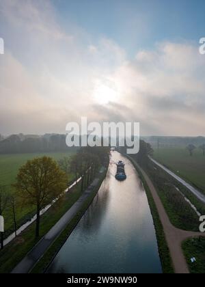 Dans cette scène aérienne tranquille, une barge descend lentement une rivière sinueuse, flanquée de rangées d'arbres dans l'air frais du matin. Le soleil, jetant un regard à travers la douce brume matinale, projette une lueur chaude et crée un chemin radieux à travers la surface de l'eau. Les champs environnants sont luxuriants et verdoyants, indiquant la richesse du paysage rural. L'image capture l'essence d'un voyage paisible à la pause de la journée, où la nature et l'activité humaine coexistent en harmonie. Voyage sur la rivière Misty à l'aube avec le soleil jetant un coup d'œil. Photo de haute qualité Banque D'Images