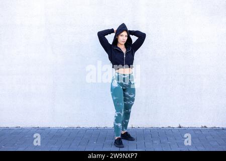 Un portrait d'une jeune femme caucasienne belle et en bonne santé portant un sweat à capuche noir, fléchissant avec confiance les muscles des deux bras avec ses mains derrière sa tête sur un fond blanc propre. Solide jeune femme Flexing muscles en sweat à capuche. Photo de haute qualité Banque D'Images