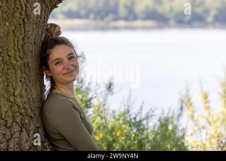 Une jeune femme millénaire brune trouve la sérénité dans l'étreinte de la nature. Elle est assise au bord d'un lac forestier, appuyée contre un arbre, profitant de la tranquillité de l'environnement verdoyant un jour d'été. Sérénité dans la nature. Photo de haute qualité Banque D'Images